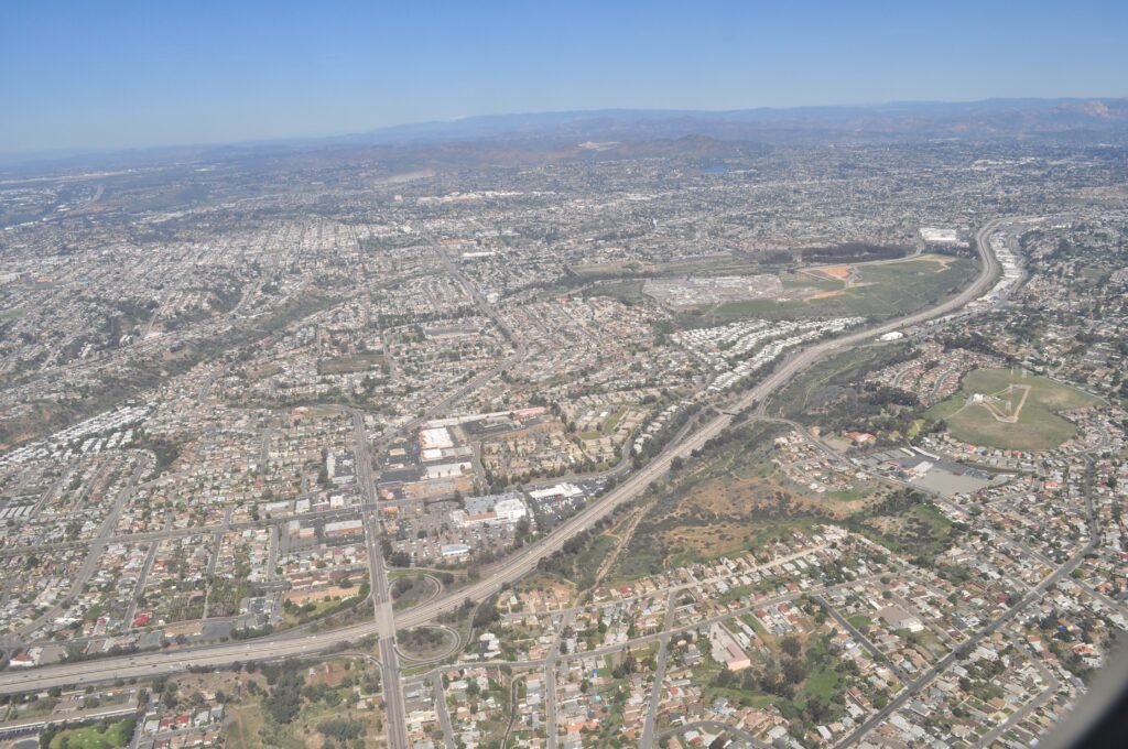 Aerial view of a densely populated suburban area in San Diego, California, featuring highways, residential neighborhoods, green spaces, and distant mountains under a clear blue sky.