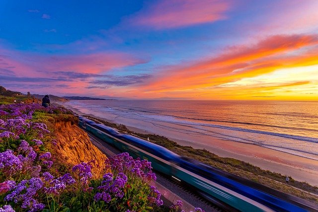 Vibrant sunset over a sandy beach with coastal cliffs, purple flowers, and an oceanfront train.