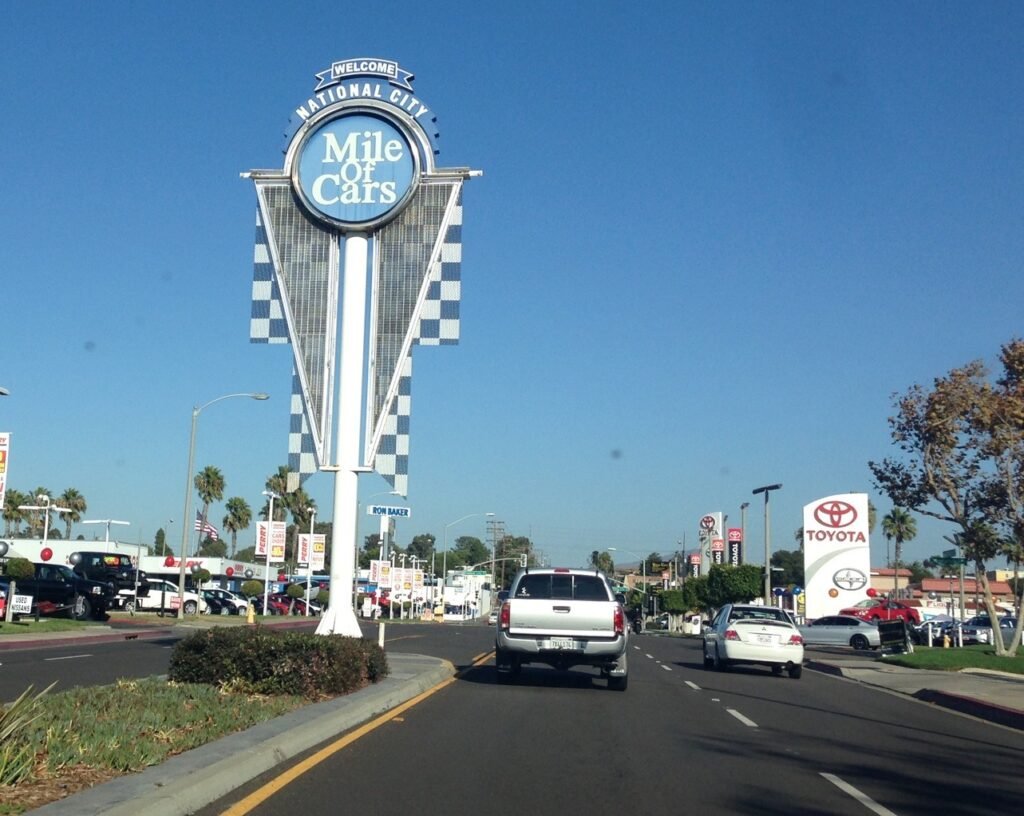 Large &ldquo;Mile of Cars&rdquo; welcome sign along a busy road lined with car dealerships in National City, California under a clear blue sky.
