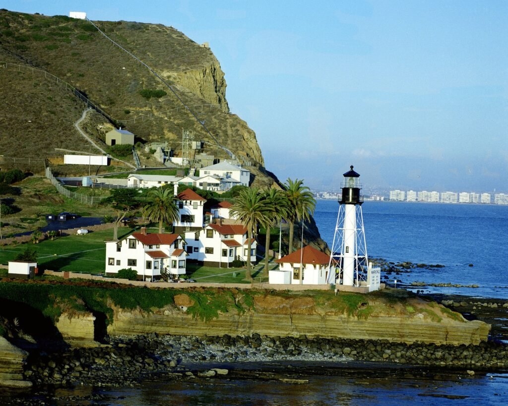 Historic lighthouse overlooking the ocean with coastal cliffs, palm trees, and seaside buildings.