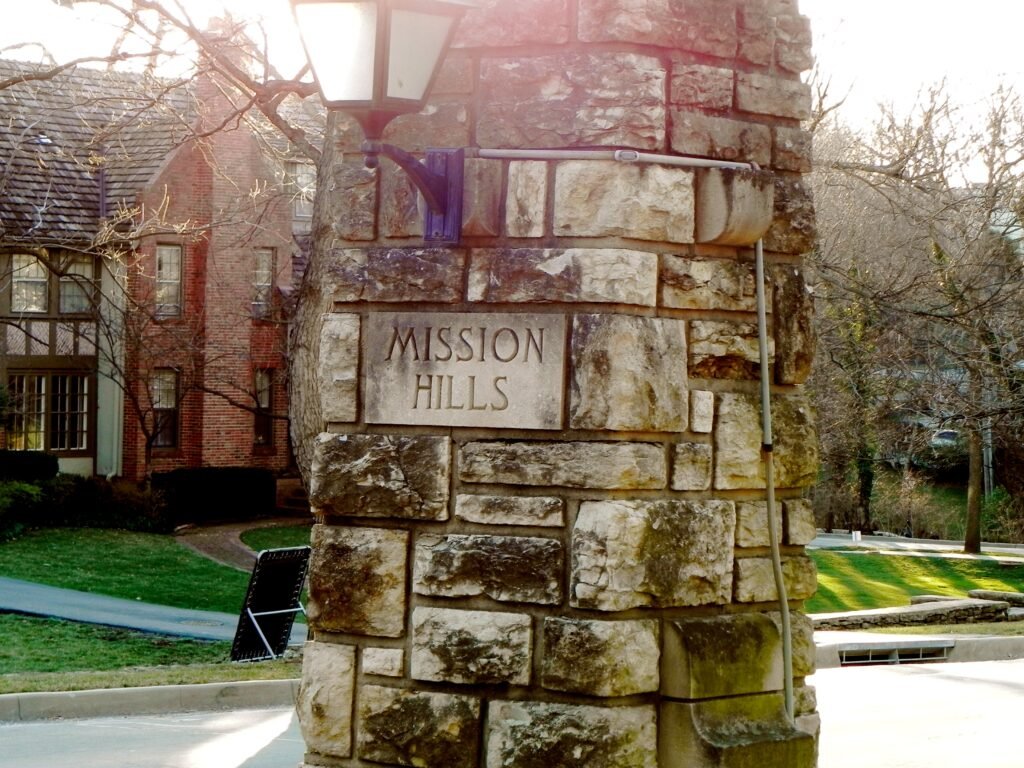 Stone entrance pillar with &ldquo;Mission Hills&rdquo; engraved on it beside a residential street and historic-style homes.