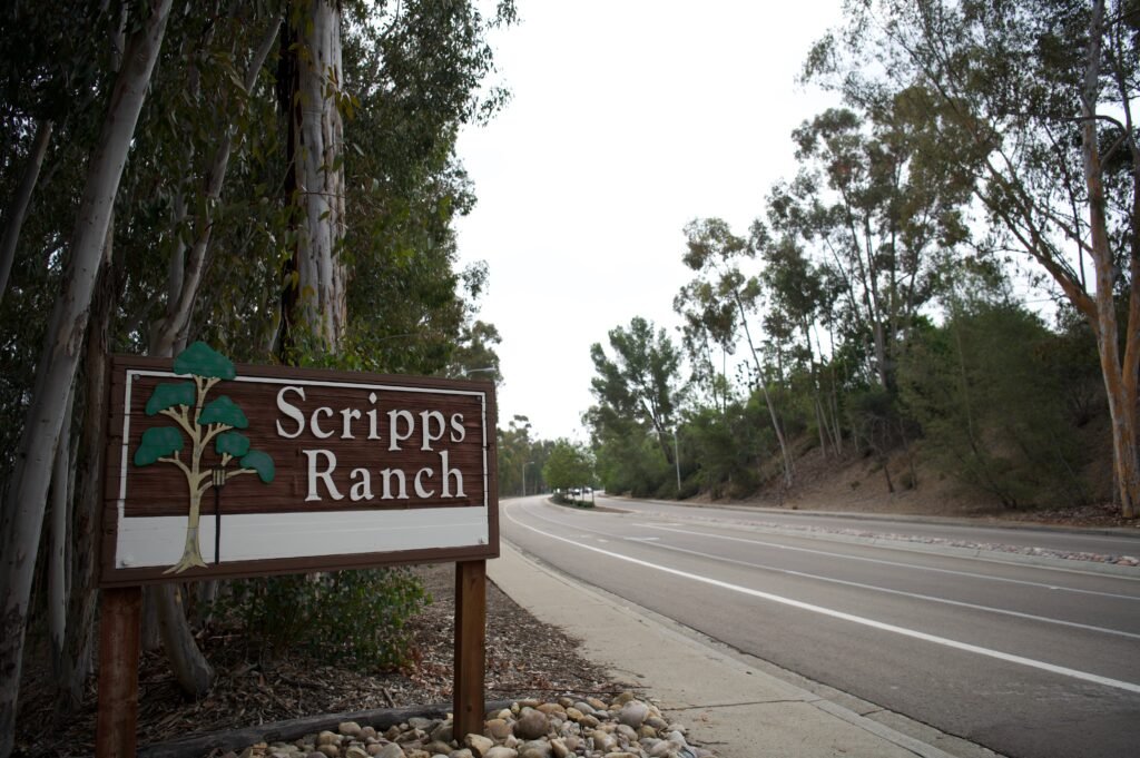 Wooden Scripps Ranch neighborhood sign beside a quiet road lined with tall trees and natural landscape.