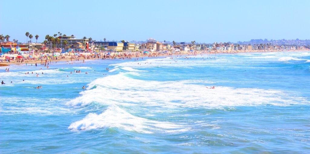 Crowded Pacific Beach shoreline in San Diego with rolling blue ocean waves, surfers, palm trees, and sunny coastal homes under a clear summer sky.