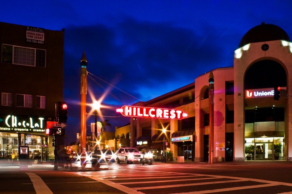 Nighttime intersection in San Diego&rsquo;s Hillcrest neighborhood with a glowing red Hillcrest sign, traffic lights, and passing cars.