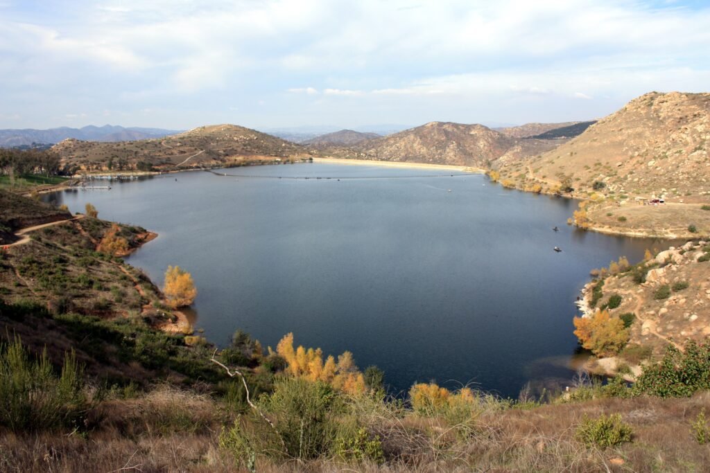 Wide view of Lake Poway surrounded by rolling hills, dry mountain terrain, and calm blue water under a cloudy sky.