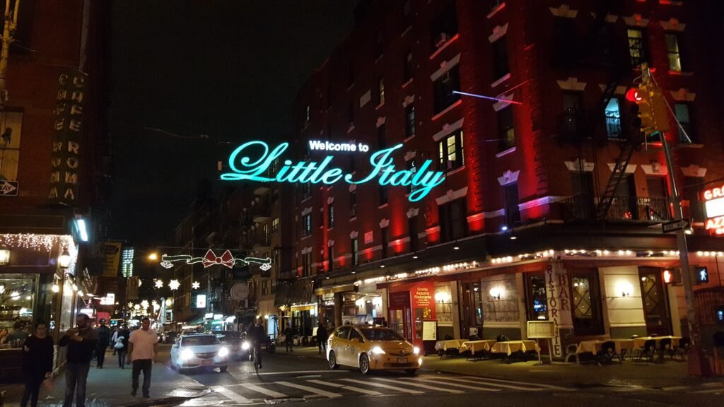 Night view of New York&rsquo;s Little Italy with glowing neon signage, busy streets, yellow taxis, and illuminated restaurants.