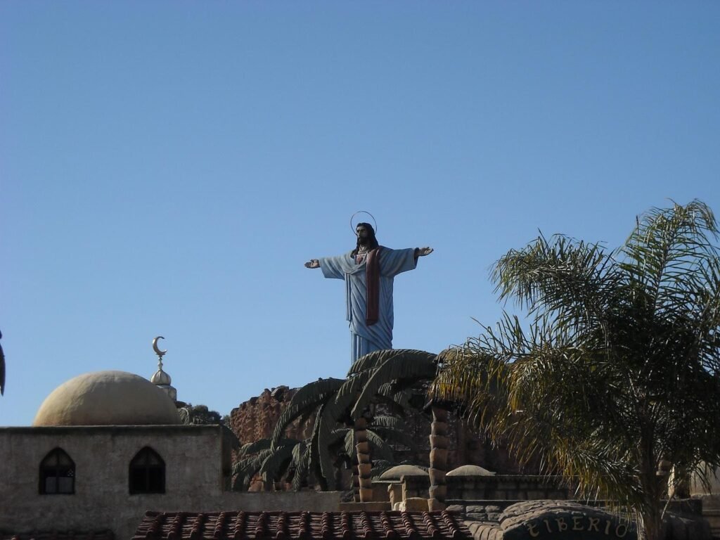 Large statue of Jesus with open arms above palm trees and domed buildings under a clear blue sky.