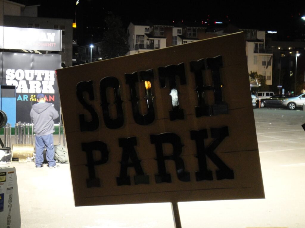 Nighttime view of a large &ldquo;South Park&rdquo; sign being dismantled in a parking lot, with city buildings and lights in the background.