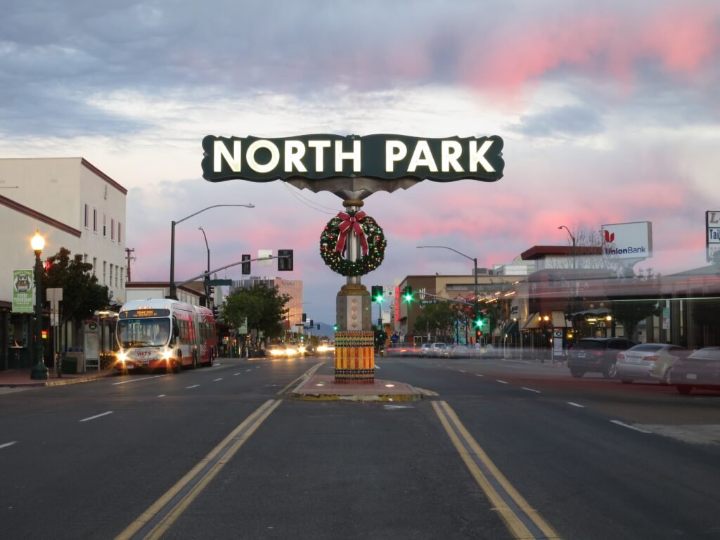 Illuminated North Park sign above a city street with traffic, buildings, and colorful evening clouds in San Diego.